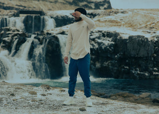 Man in een lang crèmekleurig shirt en een blauwe broek bij een waterval in IJsland